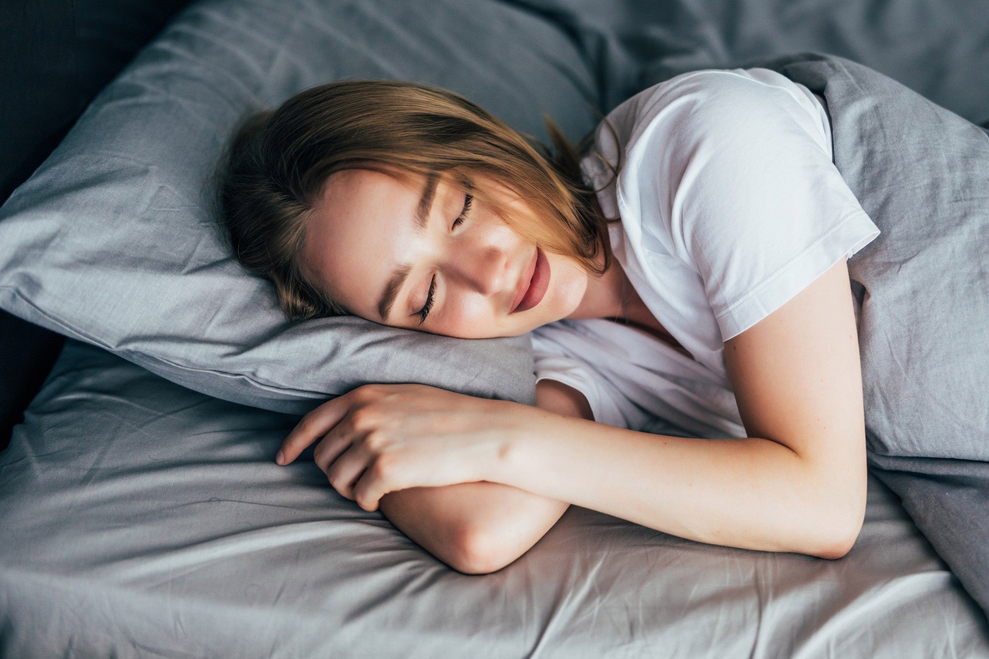 A person with long hair is sleeping on their side in bed, resting their head on a grey pillow and covered with a grey blanket. They are wearing a white shirt.